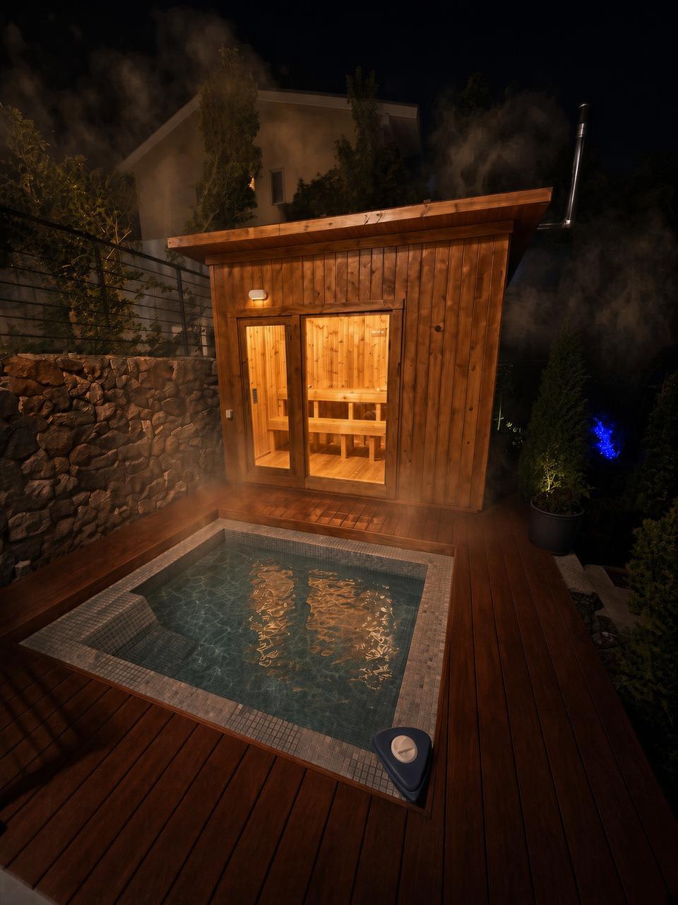Wood-burning sauna cabin glowing at night beside the steaming cold jacuzzi