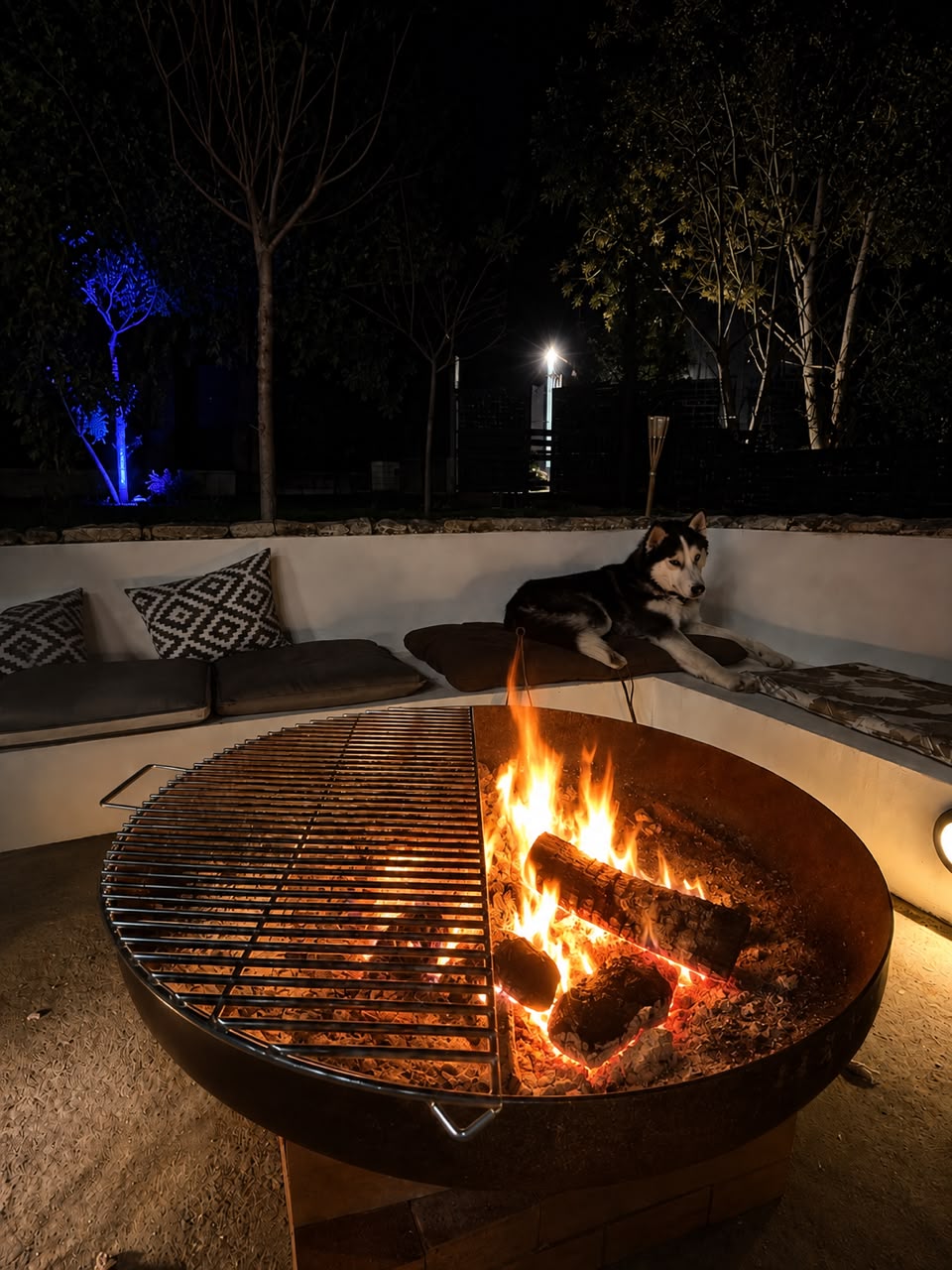Corten fire pit with a live fire and grill, ringed by sunken cushioned seating at night